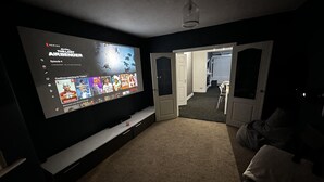 A dark room with a large TV mounted on the wall displaying a Netflix interface. There is a doorway leading to another room with a dining area visible. - 3 bed House in Blackpool With Cinema & hot tub (Blackpool)