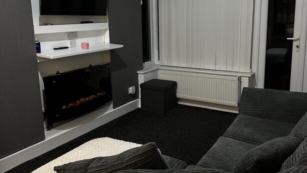 A modern living room with a large flat-screen TV mounted on a white shelf, a dark-colored sofa, and a window with white blinds. - 3 bed House in Blackpool With Cinema & hot tub (Blackpool)