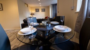 Un comedor con una mesa de vidrio dispuesta para cuatro personas, rodeada de sillas. En el fondo, un sofá y una lámpara.