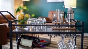Una mesa con botellas de vino y copas, una lámpara y un frutero en una habitación con paredes color turquesa y un sofá marrón.