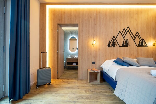 Una habitación de hotel moderna con suelo de madera, una cama con cabecera azul, una maleta y una pared de madera con diseño de montaña.