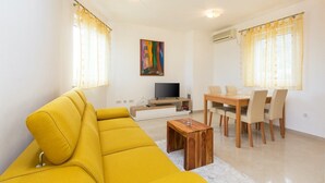 A modern living room with a yellow sofa, a wooden coffee table, a TV, and a dining area with chairs and a table.