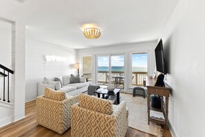 A living room with a sofa, armchairs, a coffee table, a TV, and a balcony with a view of the sea.