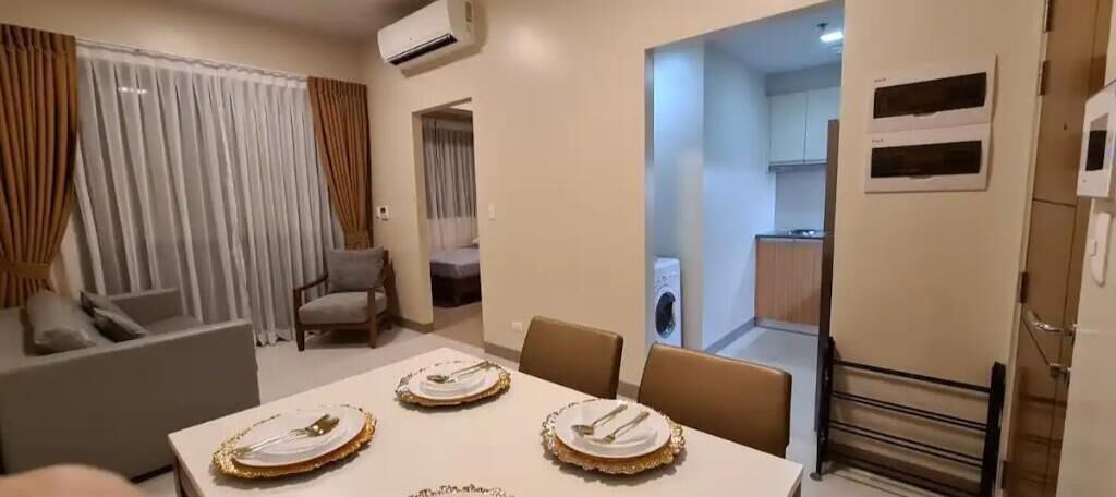 Comedor con mesa lista para cuatro personas, un sofá y una silla. Se ve una cocina a través de un arco abierto.