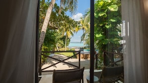 Vue depuis une chambre avec balcon donnant sur une plage, avec une piscine, des chaises et une végétation tropicale.