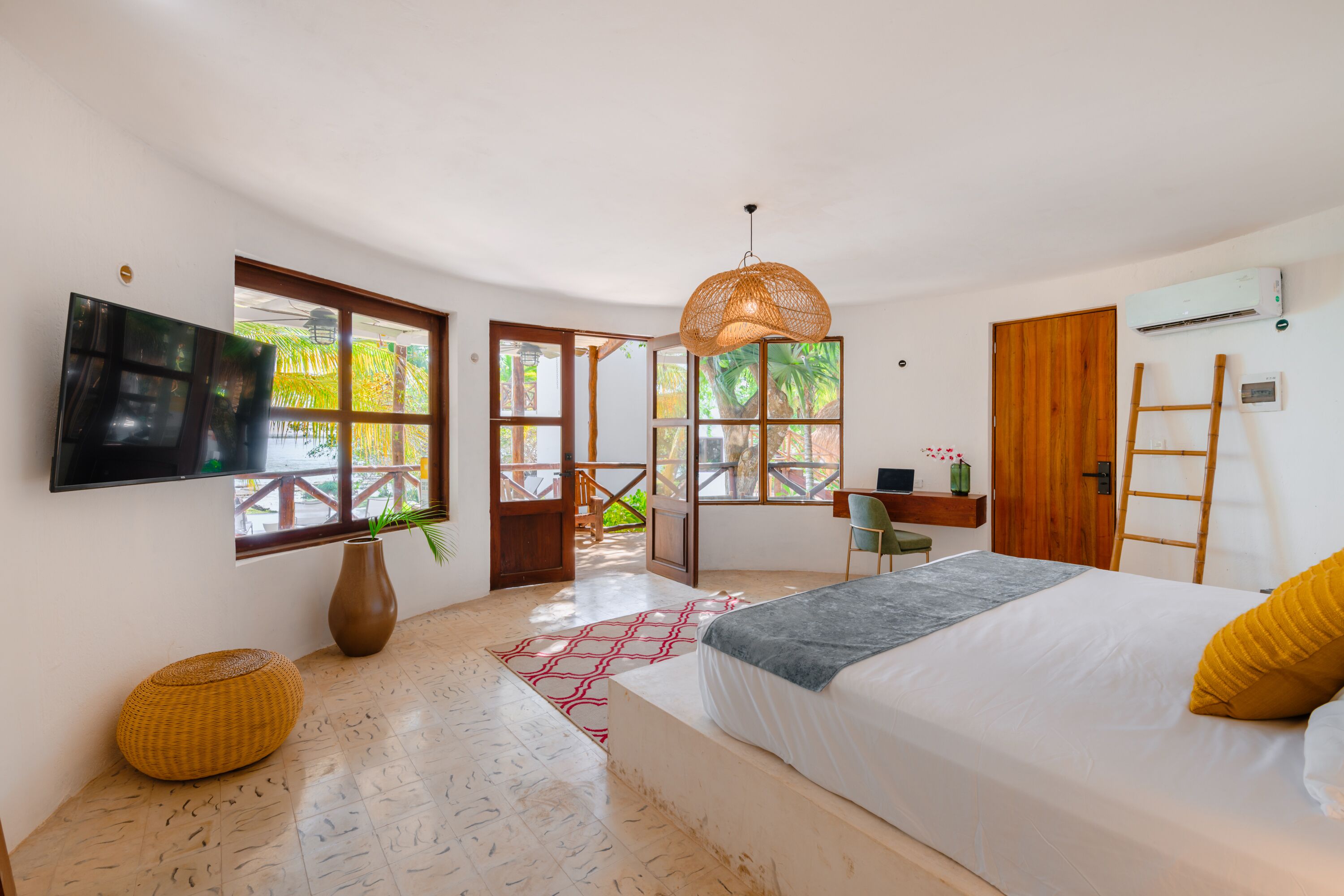 A modern bedroom with a large bed, a TV mounted on the wall, a wooden ladder, and a view of the outdoors through large windows.