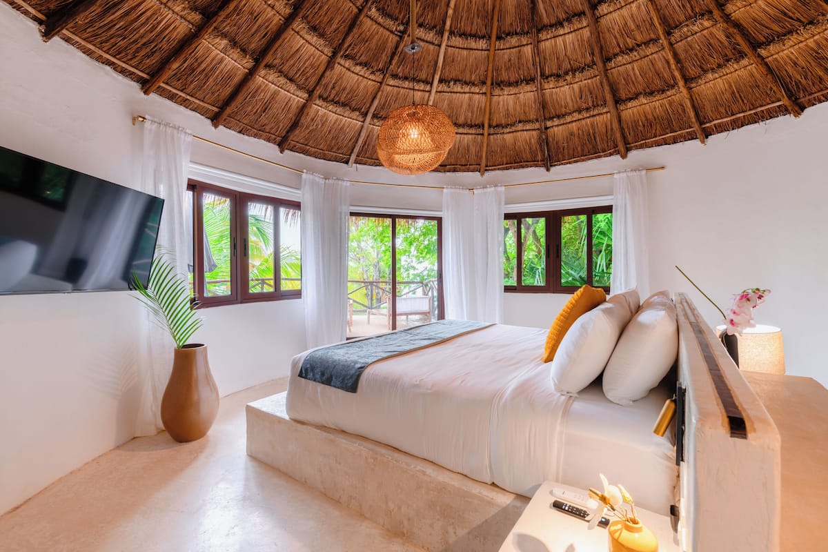 A bedroom with a large bed, a TV mounted on the wall, a window with curtains, and a thatched roof.