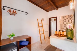 Un dormitorio con techo de madera, una cama, una mesita de noche con artículos de aseo, un estante con una planta, una escalera y un baño con baldosas estampadas.