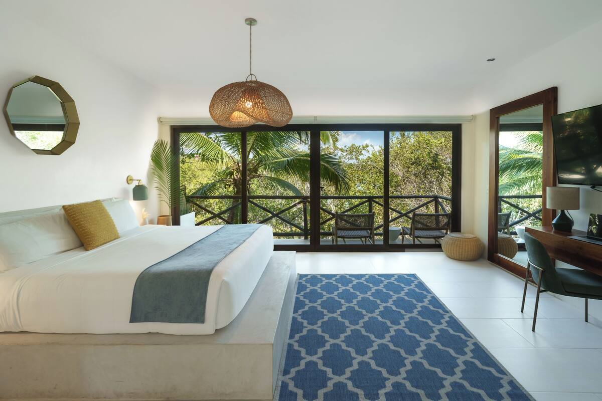 A bedroom with a large bed, a blue patterned rug, a hanging light fixture, and a view of palm trees through large windows.