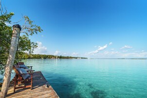 A wooden dock with two chairs and a table, overlooking clear turquoise water with a distant island and sailboats.