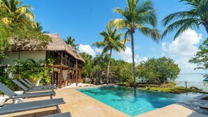 Un área junto a la piscina con sillones, una estructura de techo de paja y palmeras.