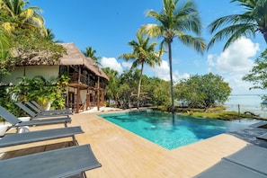 Un área junto a la piscina con sillones, una estructura de techo de paja y palmeras.