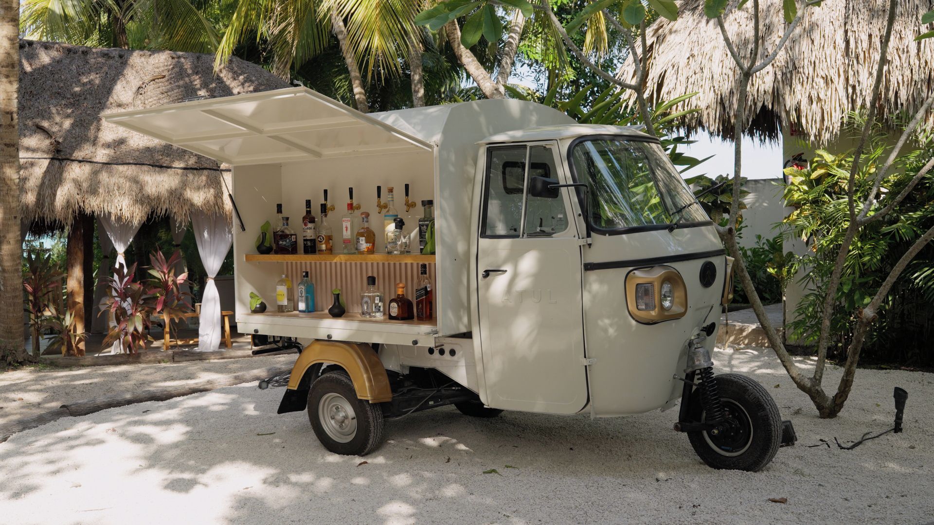 A small, white, three-wheeled vehicle with a covered cargo area containing various bottles, parked on a sandy surface with palm trees and a thatched-roof structure in the background.