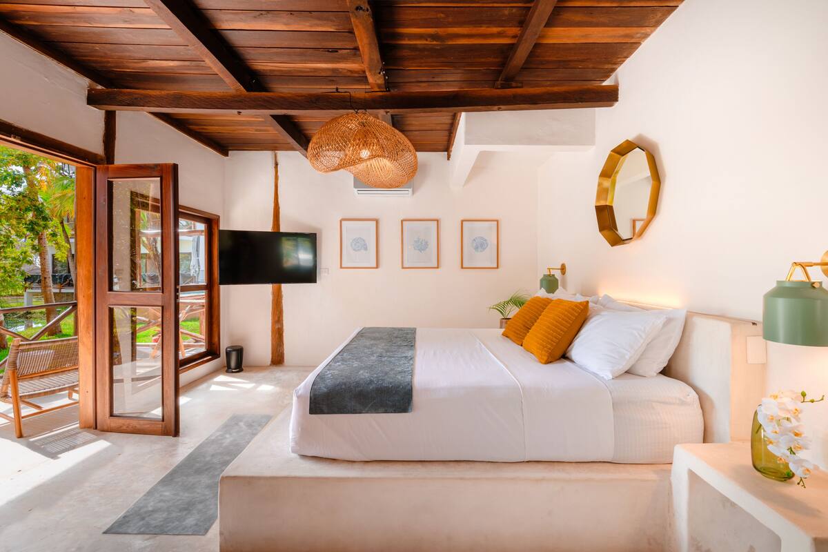 A bedroom with a bed, a TV, framed pictures, a mirror, and a window with a view of greenery.