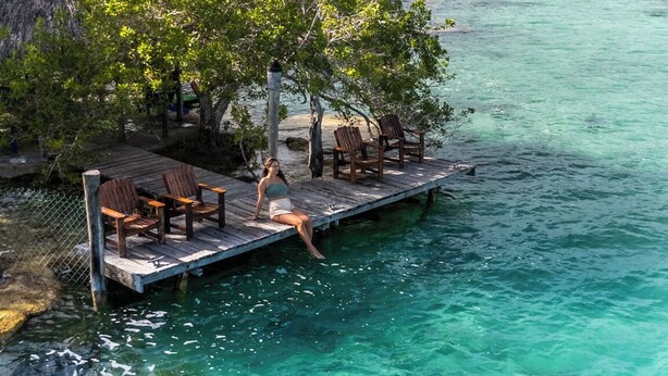 Une jetée en bois s’étendant dans une eau turquoise et limpide, avec une personne assise sur la jetée.