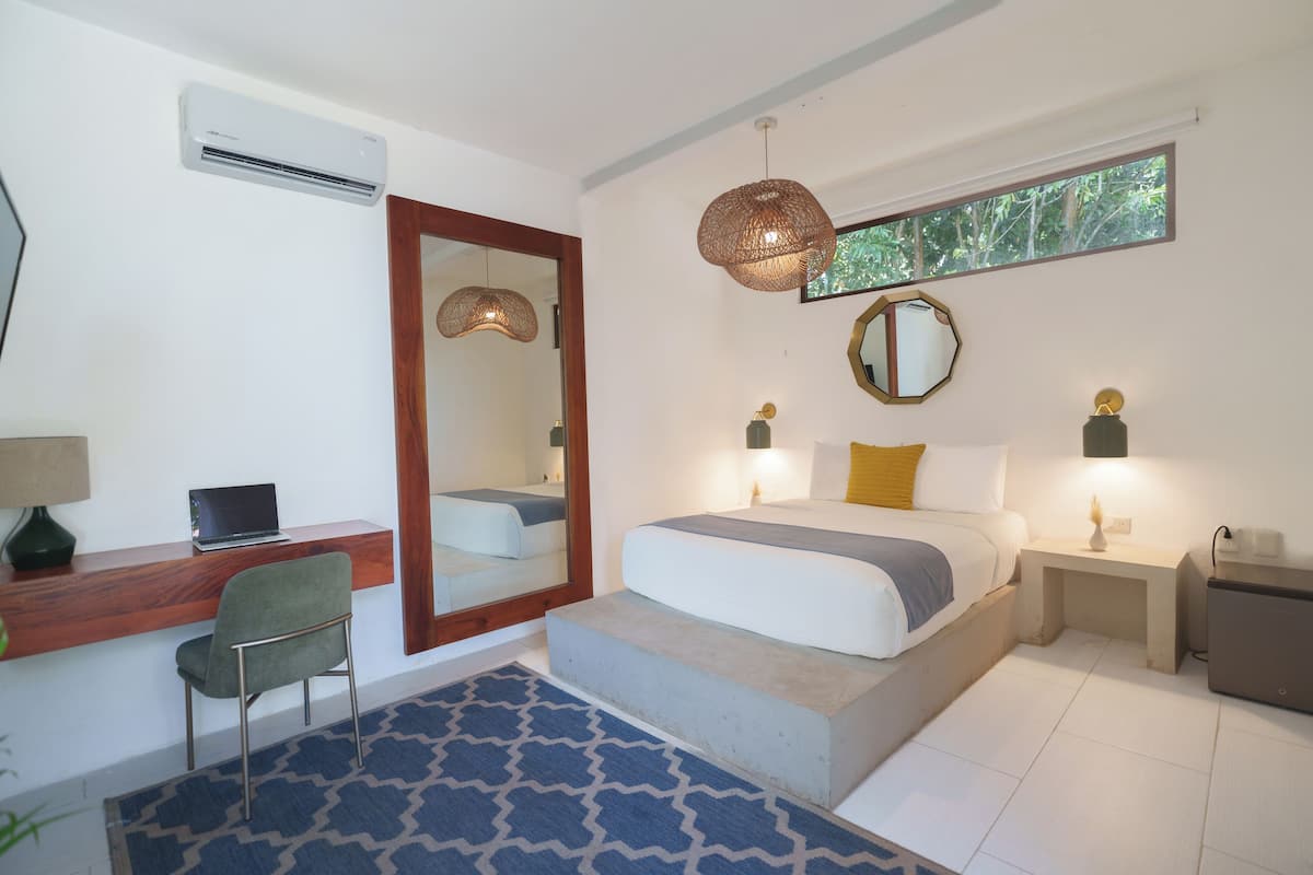 A bedroom with a bed, bedside tables, a desk with a chair, a mirror, and an air conditioning unit.