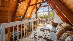 A cozy attic bedroom with a sloped wooden ceiling, a cowhide rug, and a view of the outdoors through large windows.