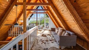 A cozy attic room with a wooden ceiling, a cowhide rug, a gray sofa, and a view of the outdoors through large windows.