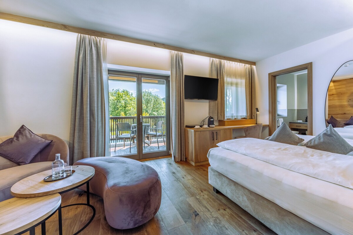 Ett hotellrum med en säng, en soffa, ett litet bord, en TV och en balkong med utemöbler.