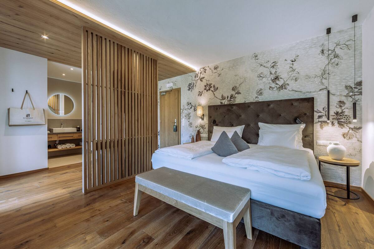 Une chambre moderne avec un plancher en bois, un grand lit doté d’une tête de lit capitonnée, un banc et une petite table de chevet sur laquelle est posé un vase.
