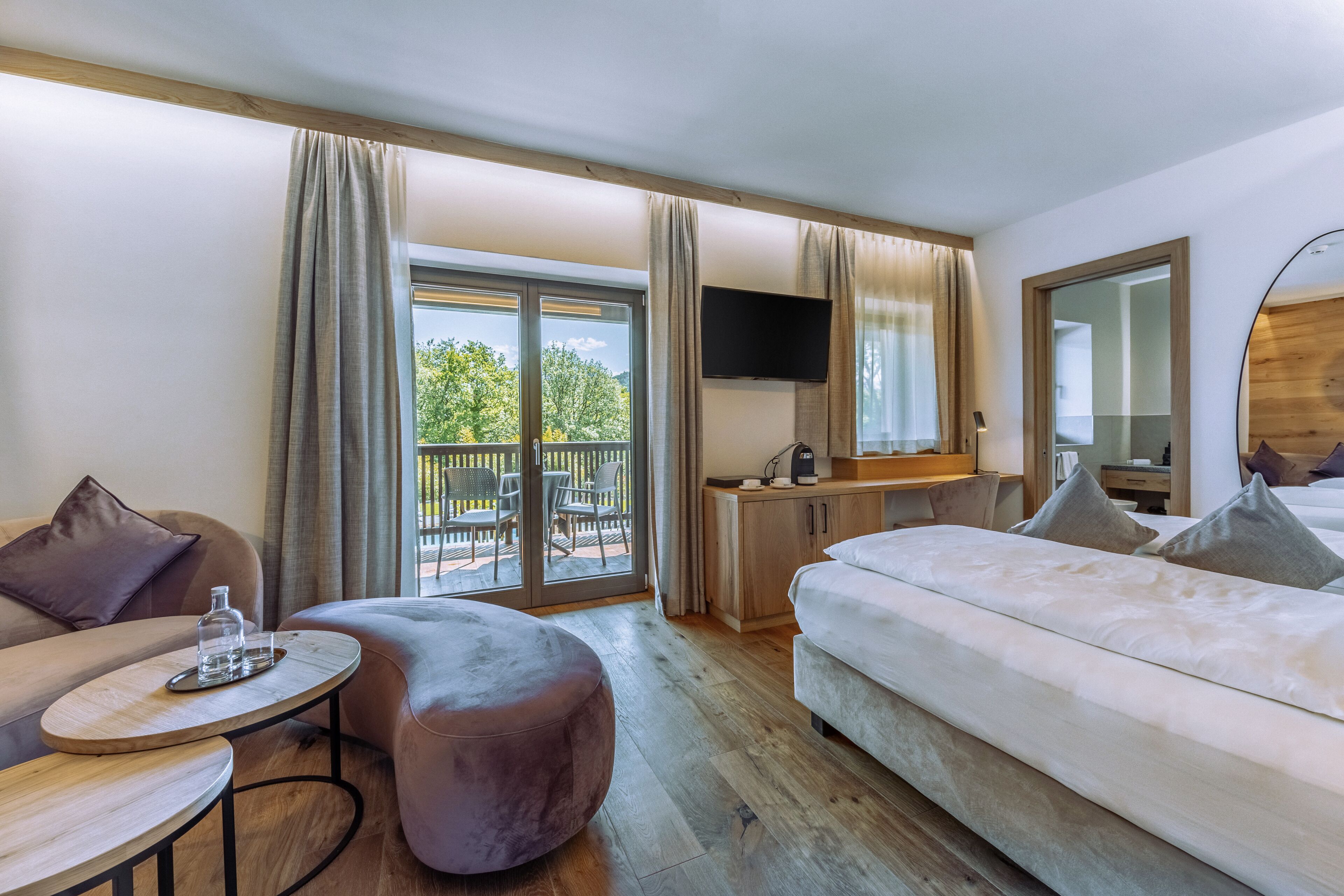 A modern hotel room with a sofa, a round ottoman, a bed, a desk, and a TV.