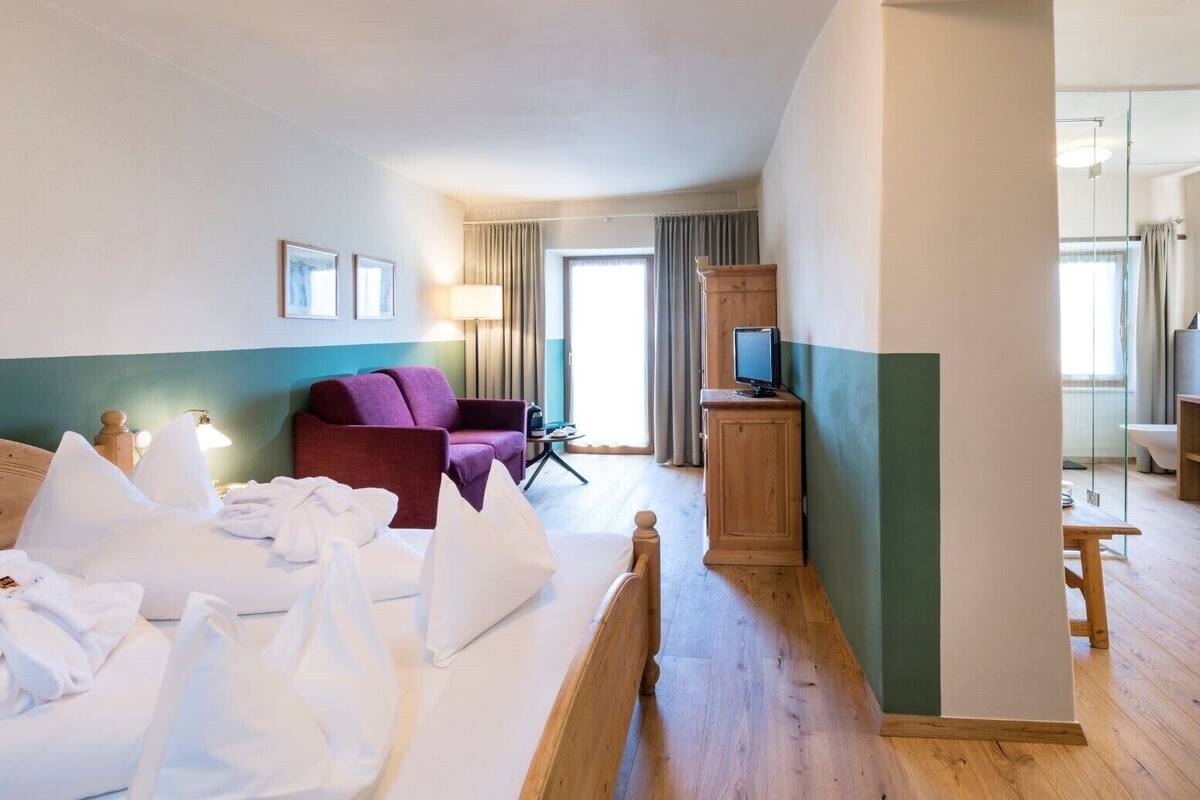 Une chambre d’hôtel avec un lit, un fauteuil violet, une armoire en bois avec un téléviseur et une fenêtre avec des rideaux.