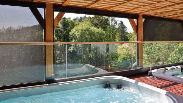 Vasca idromassaggio con terrazza in legno e struttura in vetro, immersa in una vista boschiva.