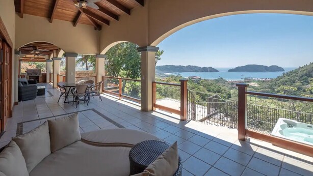 Un balcón amplio con vistas al mar y las colinas, un jacuzzi y una zona de estar con mesa y sillas.