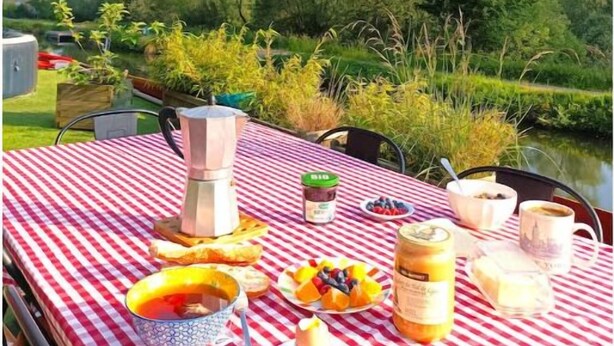 A picnic setup with a checkered tablecloth, a variety of food items, and a view of a river and trees in the background. - Exceptionnel. Bateau Logement (Ailly-sur-Somme)