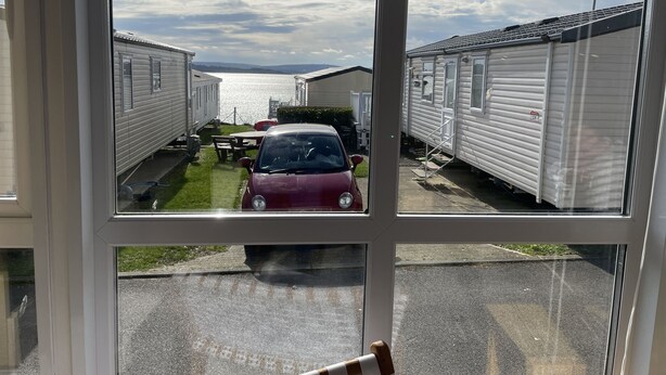 Une vue à travers la fenêtre d’un camping, avec une voiture rouge garée au premier plan, une table de pique-nique et une étendue d’eau en arrière-plan.