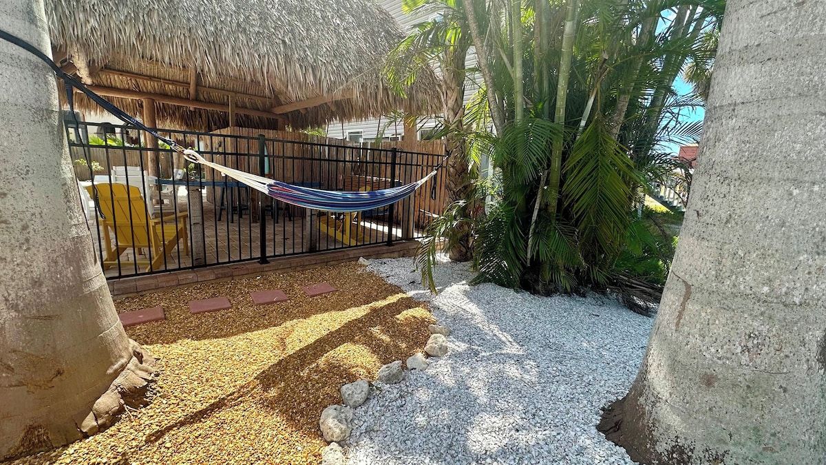 A hammock tied between two concrete pillars, a thatched-roof structure, and a palm tree.