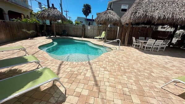 A swimming pool with a brick patio, lounge chairs, and thatched umbrellas.