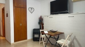 A compact living space with a wooden wardrobe, a flat-screen TV mounted on a white cabinet, a small table with chairs, and a heater. - TERRASOL PLAZAMAR 4-2 (Torre del Mar)
