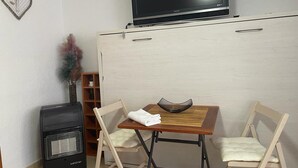 A compact living space with a flat-screen TV mounted above a wooden table and two foldable chairs. A small heater is placed against the wall, and a decorative heart-shaped item hangs on the wall. - TERRASOL PLAZAMAR 4-2 (Torre del Mar)
