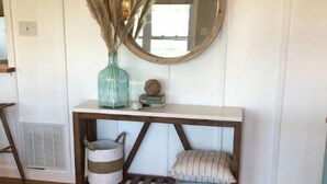 Une pièce avec un plancher en bois, un mur blanc, une console en bois surmontée d’un miroir, un vase contenant des plumes d’herbe des pampas et un petit tapis.