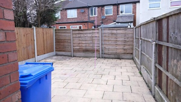 A paved backyard with a wooden fence, a blue trash bin, and a brick wall.