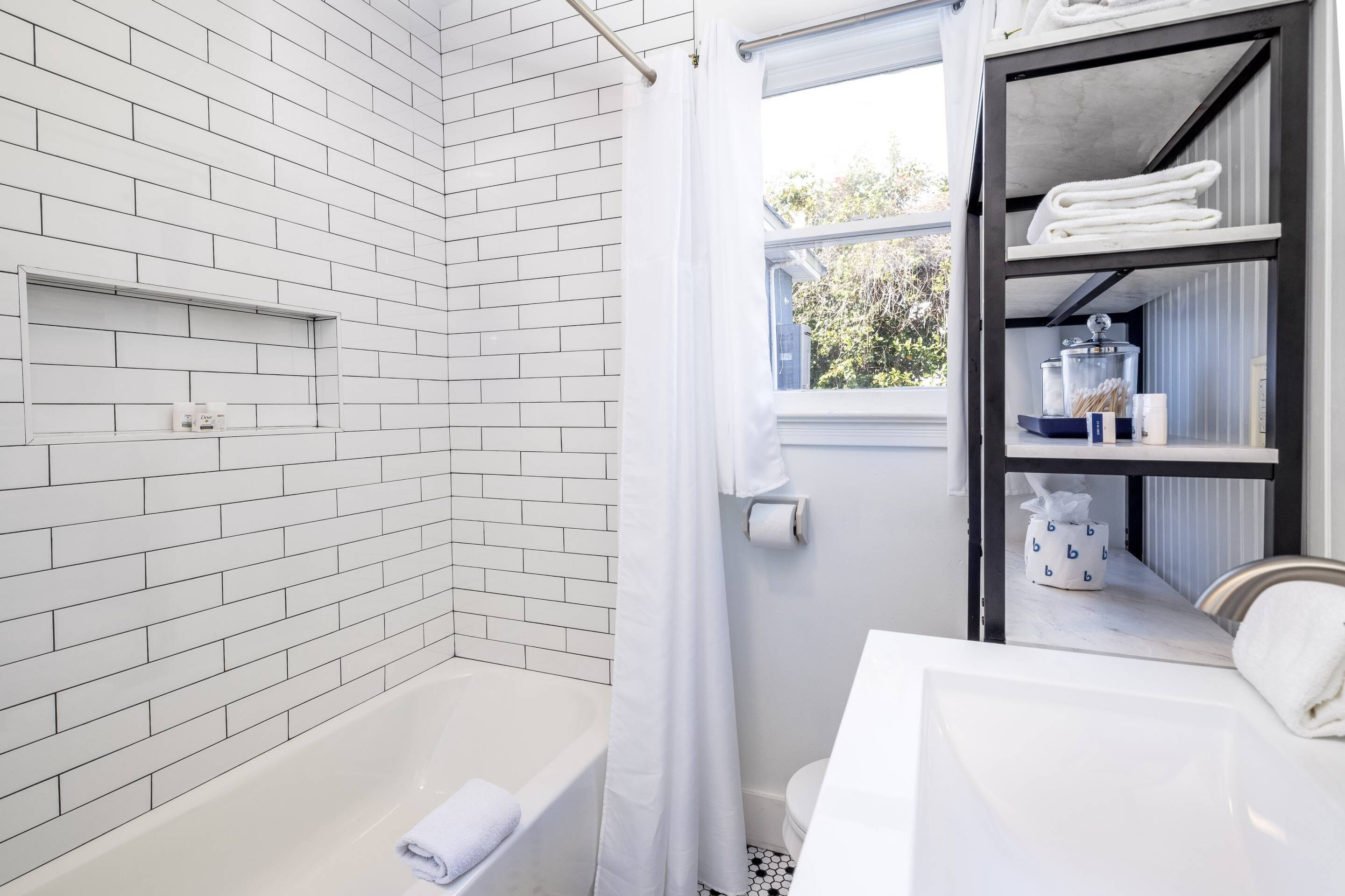 Ein modernes Badezimmer mit weißen Ziegelwänden, einer weißen Badewanne, einem Fenster mit Ausblick und einem Regal mit Handtüchern und Toilettenartikeln.