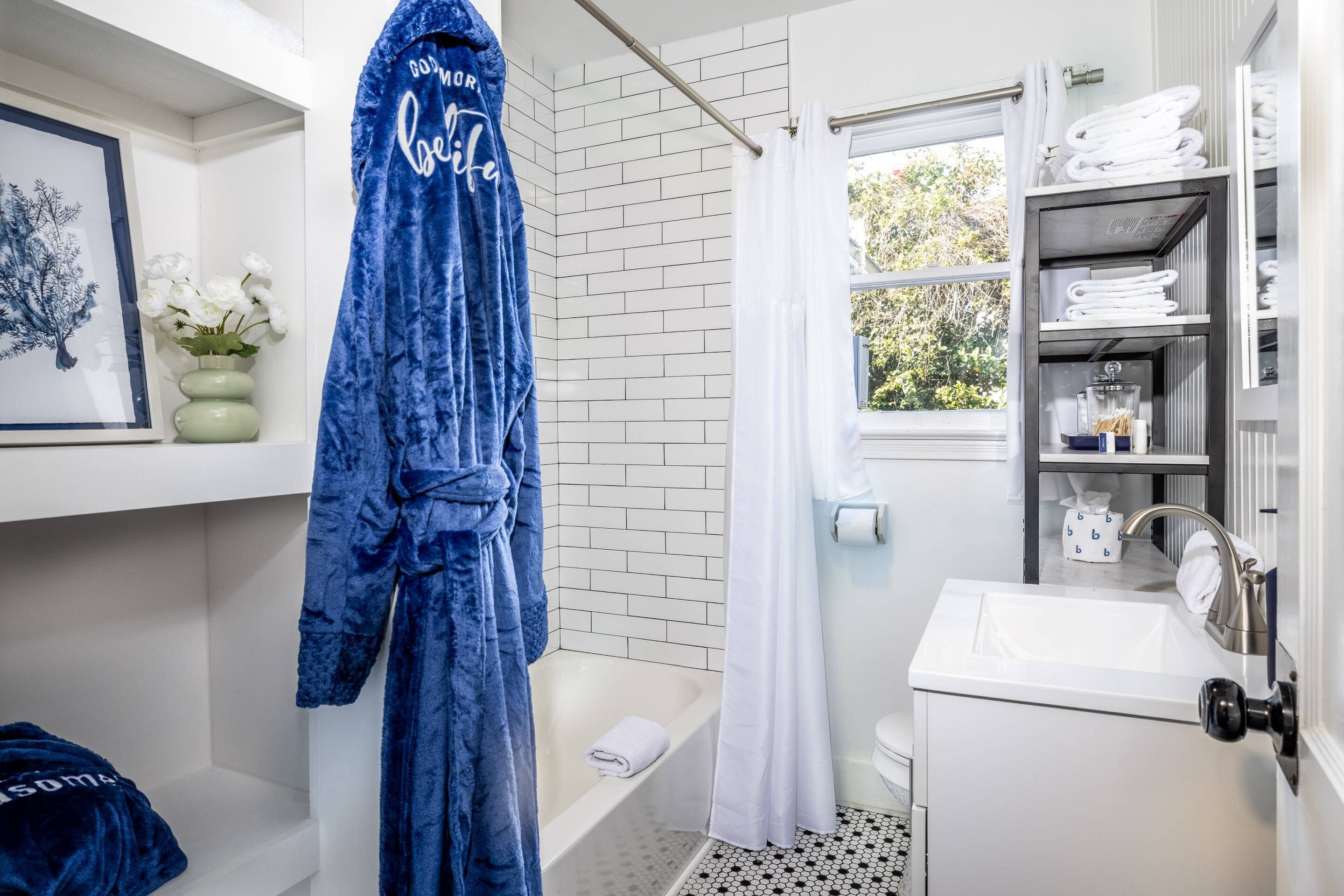 Un baño con un lavamanos blanco, una bañera y una cortina de ducha. Se ve un albornoz azul colgado en un perchero y un cuadro en la pared.
