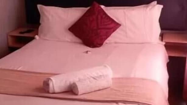 Een net opgemaakt bed met witte lakens en een rood kussen, een ingelijst beeld aan de muur, en een houten hoofdbord.