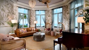 Ein geräumiges Wohnzimmer mit einem braunen Ledersofa, einem runden Couchtisch und einem Flügel. Große Fenster bieten Blick auf das Meer.