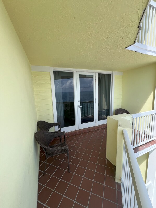 Un balcon doté d’un sol carrelé, d’une chaise en osier, d’une balustrade blanche et offrant une vue sur la mer à travers des portes vitrées.