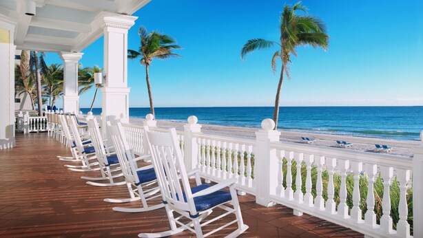 Ein Balkon am Strand mit weißen Schaukelstühlen, Palmen und Blick auf das Meer.