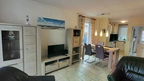 A living room with a TV, a dining area with chairs, a kitchen, and a view of the sea through a window.