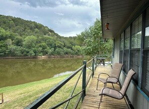 A wooden deck with chairs overlooking a lake, surrounded by lush greenery and hills.