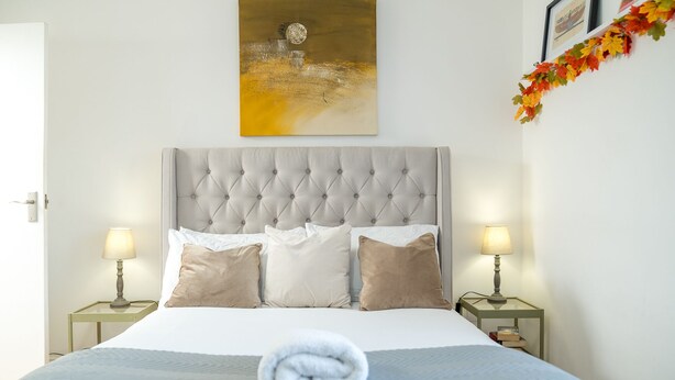 Una cama bien hecha con cabecera gris, almohadas blancas y beige, y una toalla blanca enrollada. Hay dos mesitas de noche con lámparas, un cuadro enmarcado sobre la cama y hojas de otoño en la pared.
