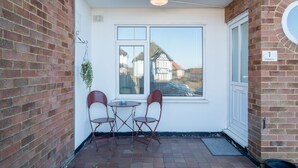 Un pequeño patio con dos sillas y una mesa, una ventana con persianas y una puerta con placa numérica.