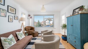 Una sala de estar moderna con un sofá marrón, dos sillones, un aparador azul y vista a una casa a través de una ventana.