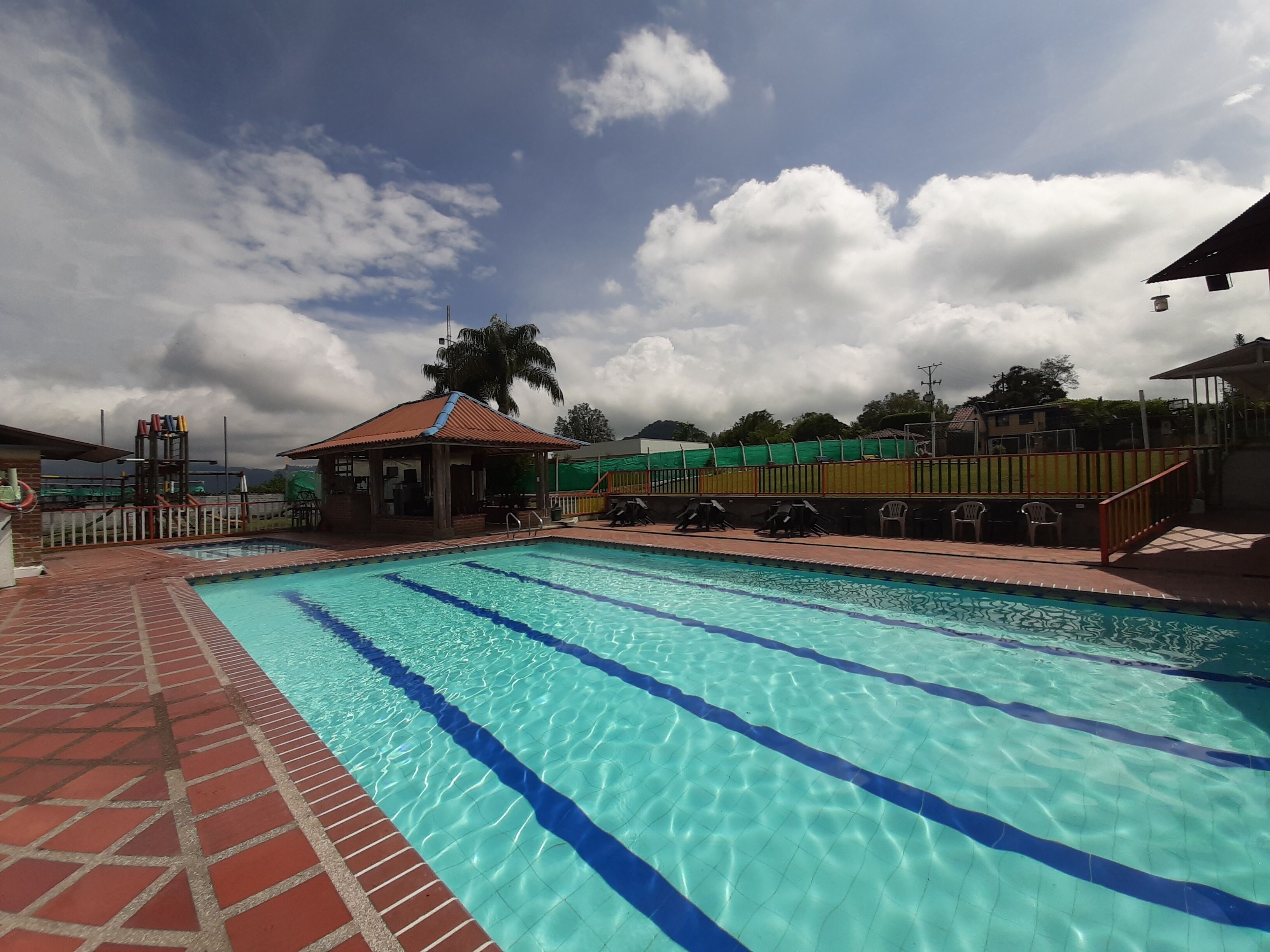 A swimming pool with clear blue water and lane dividers, surrounded by a tiled deck and a covered seating area.