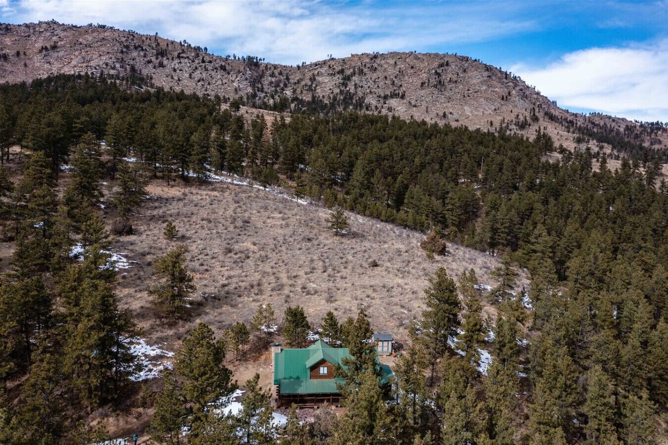 Una cabaña aislada con techo verde, ubicada entre bosques densos y un paisaje montañoso.