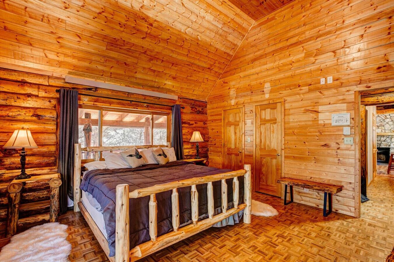 Une chambre située dans une cabane en rondins, avec un lit en bois, deux lampes de chevet, une fenêtre offrant une vue agréable et une cheminée en pierre.